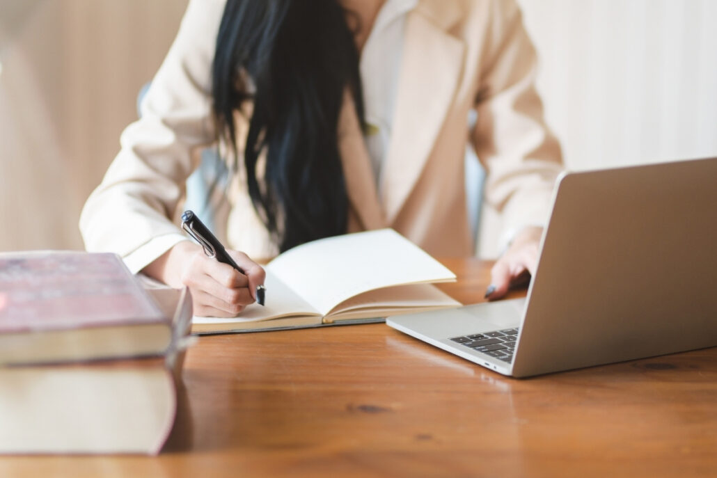 Freelancer writing in a notebook while working on a silver laptop at a desk.