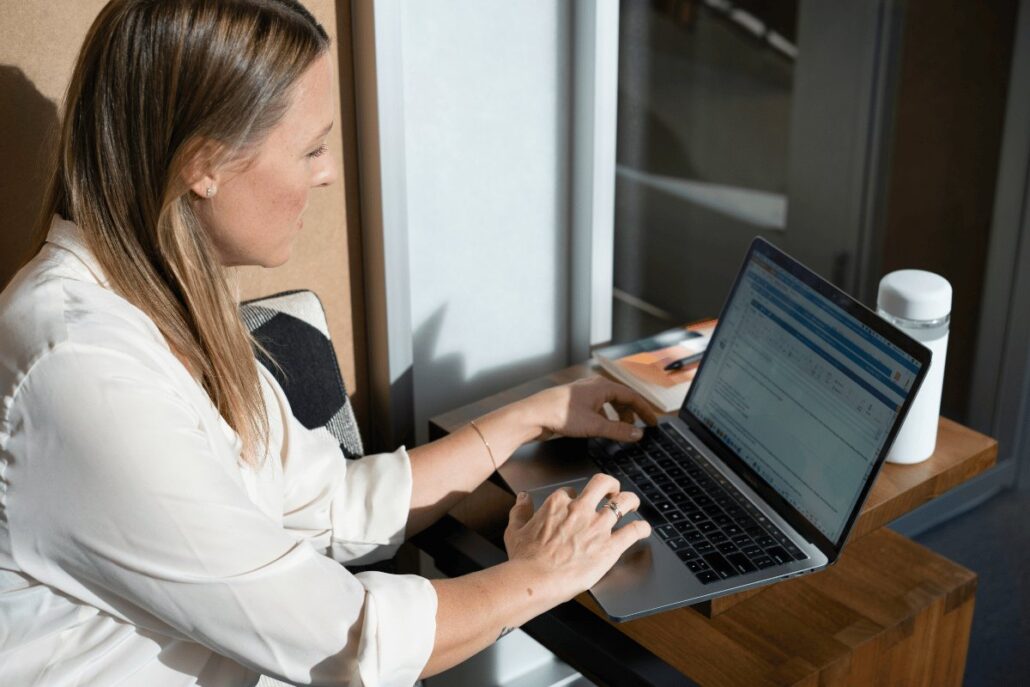 An individual sits at a wooden desk, typing on a laptop, with a notebook and a white water bottle nearby in a well-lit workspace.