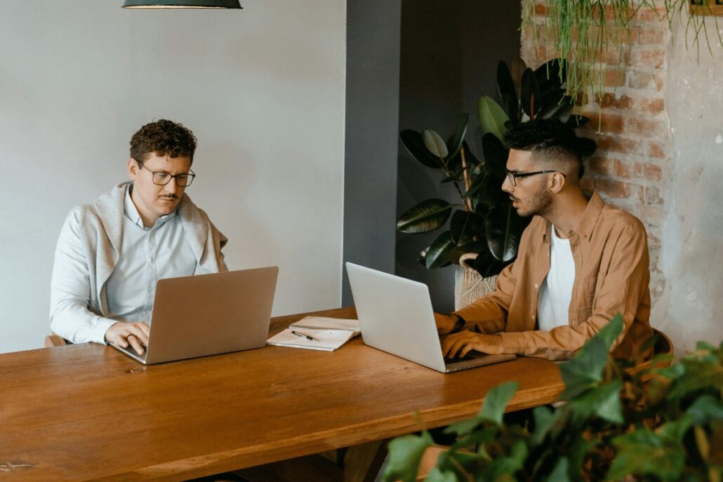Two men working on laptops at a wooden table in a modern office with plants around them.