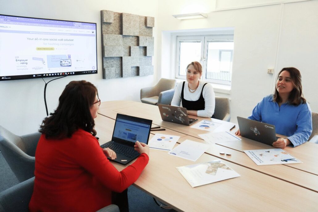 Three colleagues in a meeting room discussing a project with laptops open and a presentation on the screen.