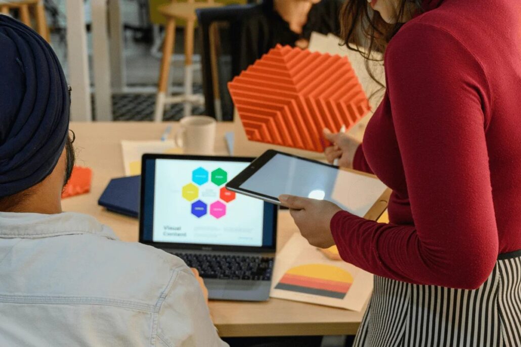 A person with a turban seated at a desk looking at a laptop while another person stands and presents a tablet.