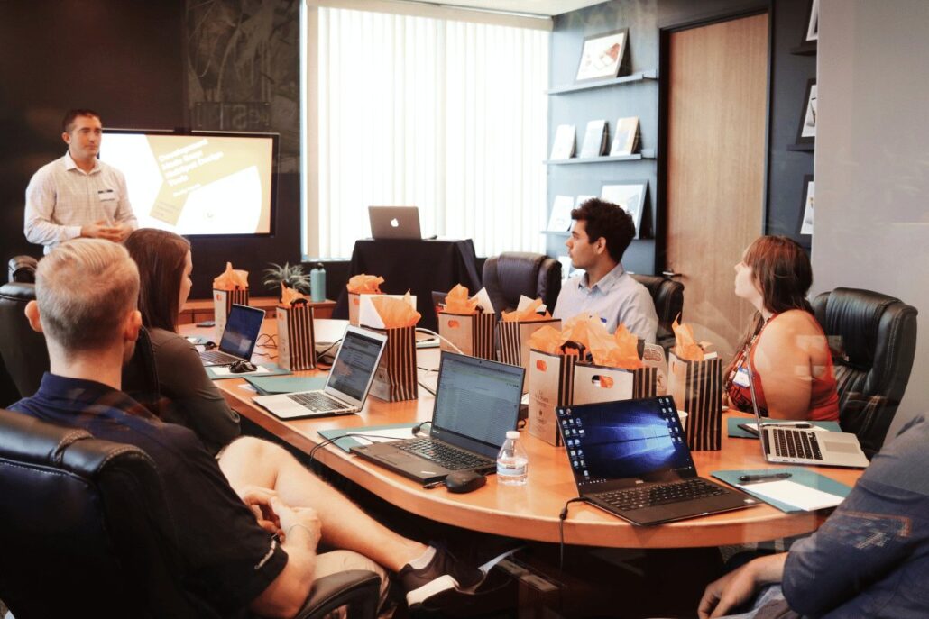 A meeting in a conference room with a presenter speaking to a group, while laptops and gift bags are on the table.