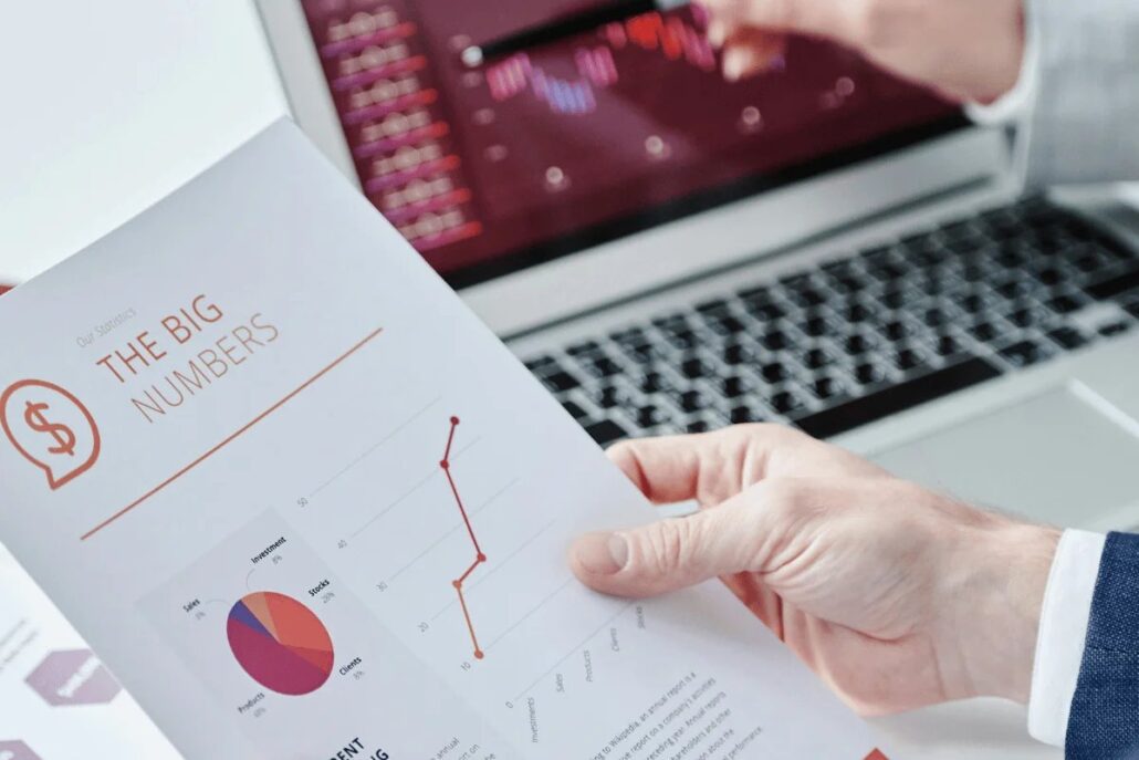 A person holding a report with a chart titled "The Big Numbers," while reviewing data on a laptop screen.