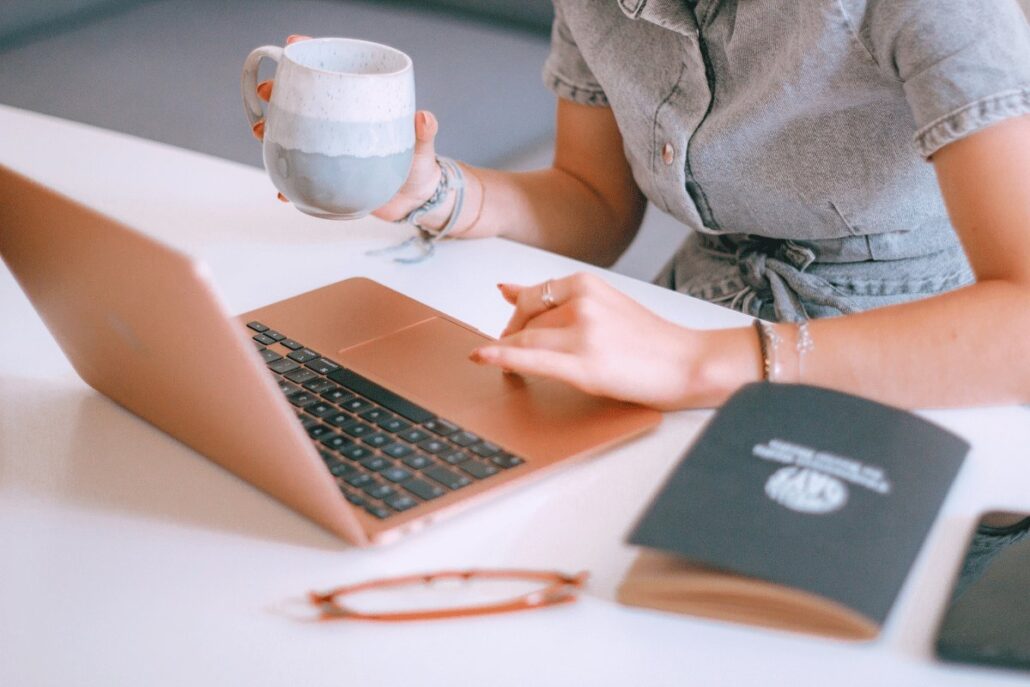 A person in a denim shirt holds a mug while typing on a rose gold laptop, with glasses and a notebook on the table.