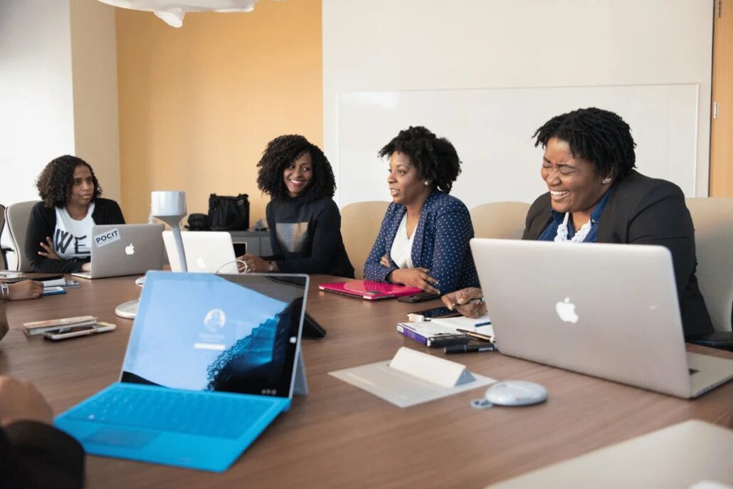 A group of four women sitting at a conference table with laptops, engaged in conversation and smiling.