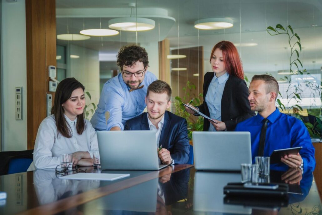 A group of professionals collaborates around laptops in a modern office setting, discussing ideas and reviewing documents.