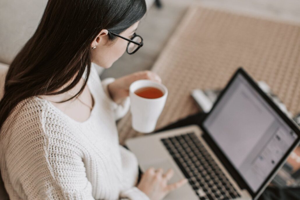 A person sitting at a desk with a laptop, holding a cup of tea while typing.