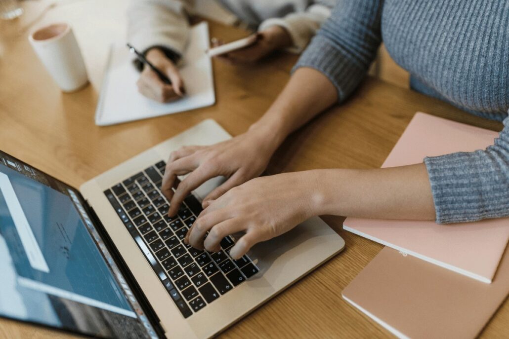 Two people working at a desk, one typing on a laptop and the other writing notes.