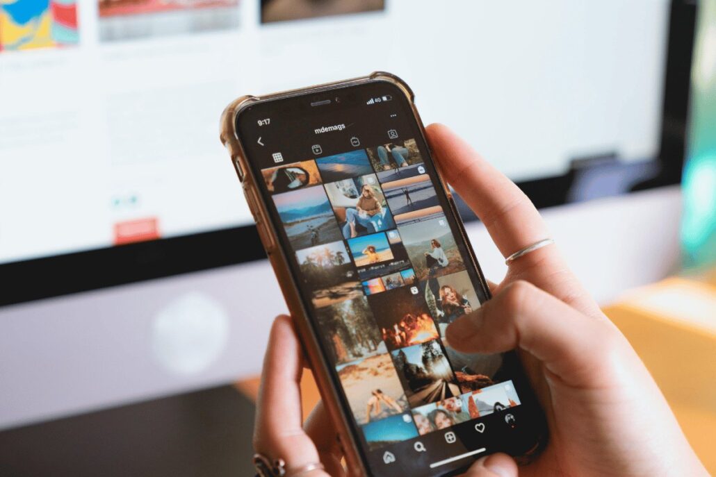 A person scrolling through Instagram on their phone, with a desktop screen in the background.