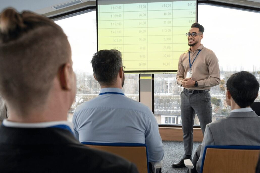 A presenter speaks in front of an audience, displaying data on a large screen in a modern meeting room with a city view.