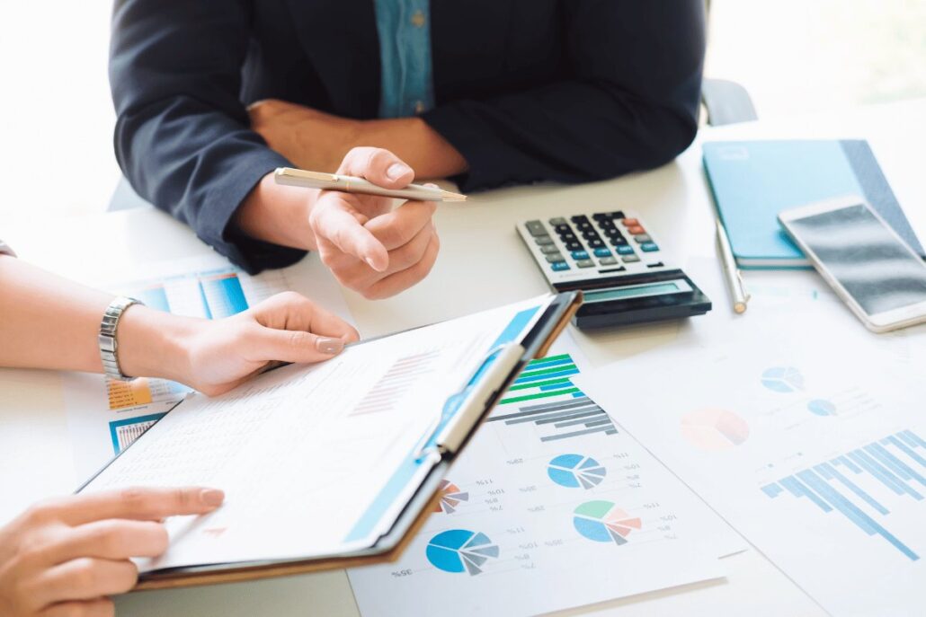 Two professionals are discussing charts and graphs on a report, with a calculator, phone, and notebooks on the table.