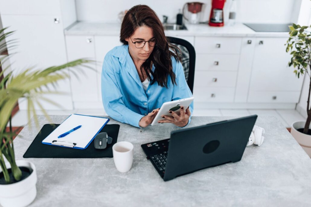 Woman in a blue shirt working on a tablet at a kitchen counter with a laptop and clipboard nearby.