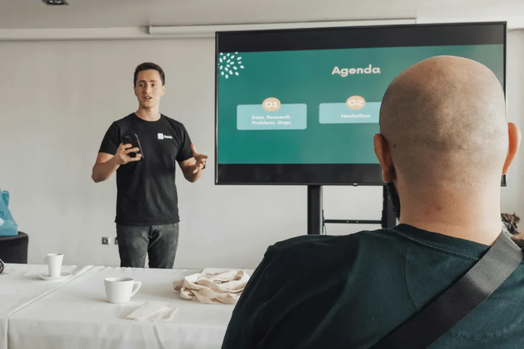 A presenter engages an audience in a modern meeting room, discussing an agenda displayed on a large screen, featuring two key topics.