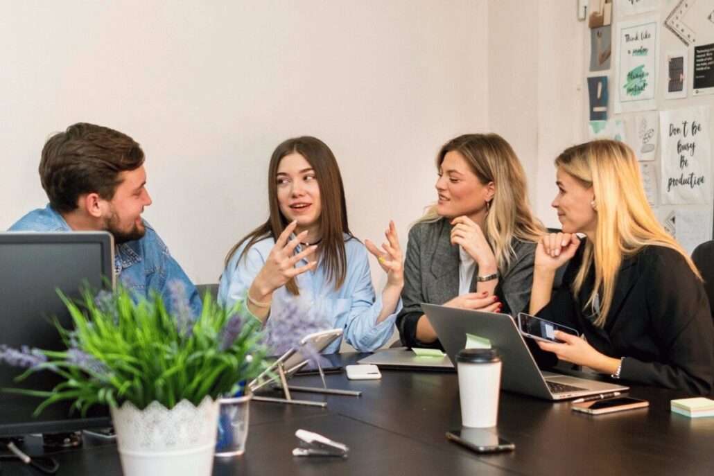 A group of four people engaged in discussion around a conference table with laptops, phones, and a potted plant.