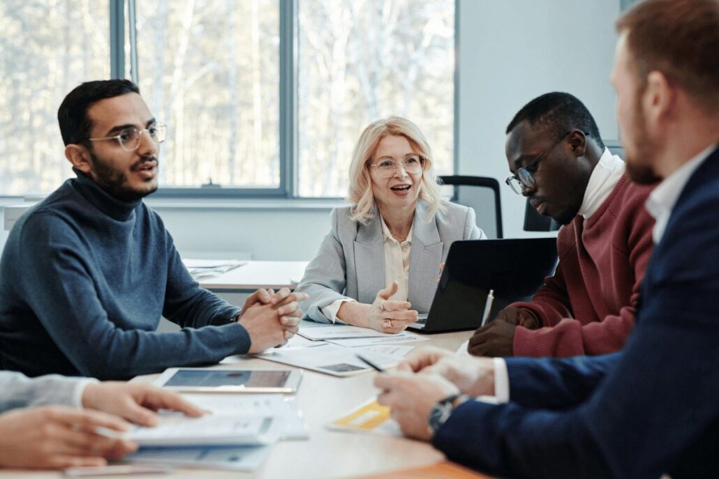 A diverse group of professionals engages in a meeting around a table, with papers and a laptop in view, discussing ideas.