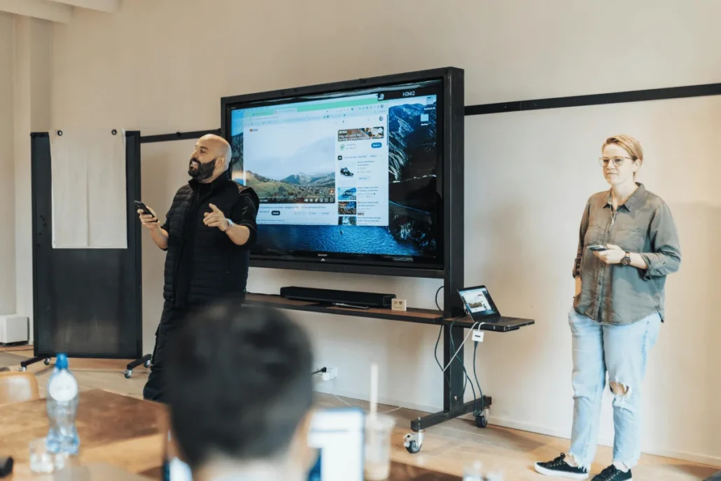 Two presenters stand in front of a large screen displaying a scenic landscape, engaging the audience while holding their phones.