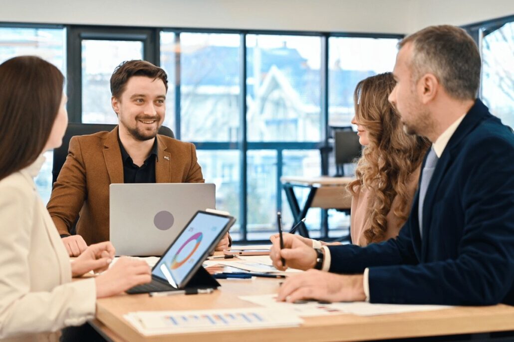 A business meeting scene with four professionals discussing documents, a laptop, and a tablet displaying a colorful graph. Bright, modern setting.