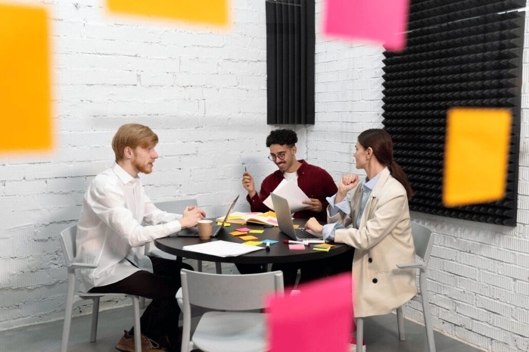 Three individuals engage in a discussion around a table cluttered with laptops, papers, and colorful sticky notes in a modern workspace.