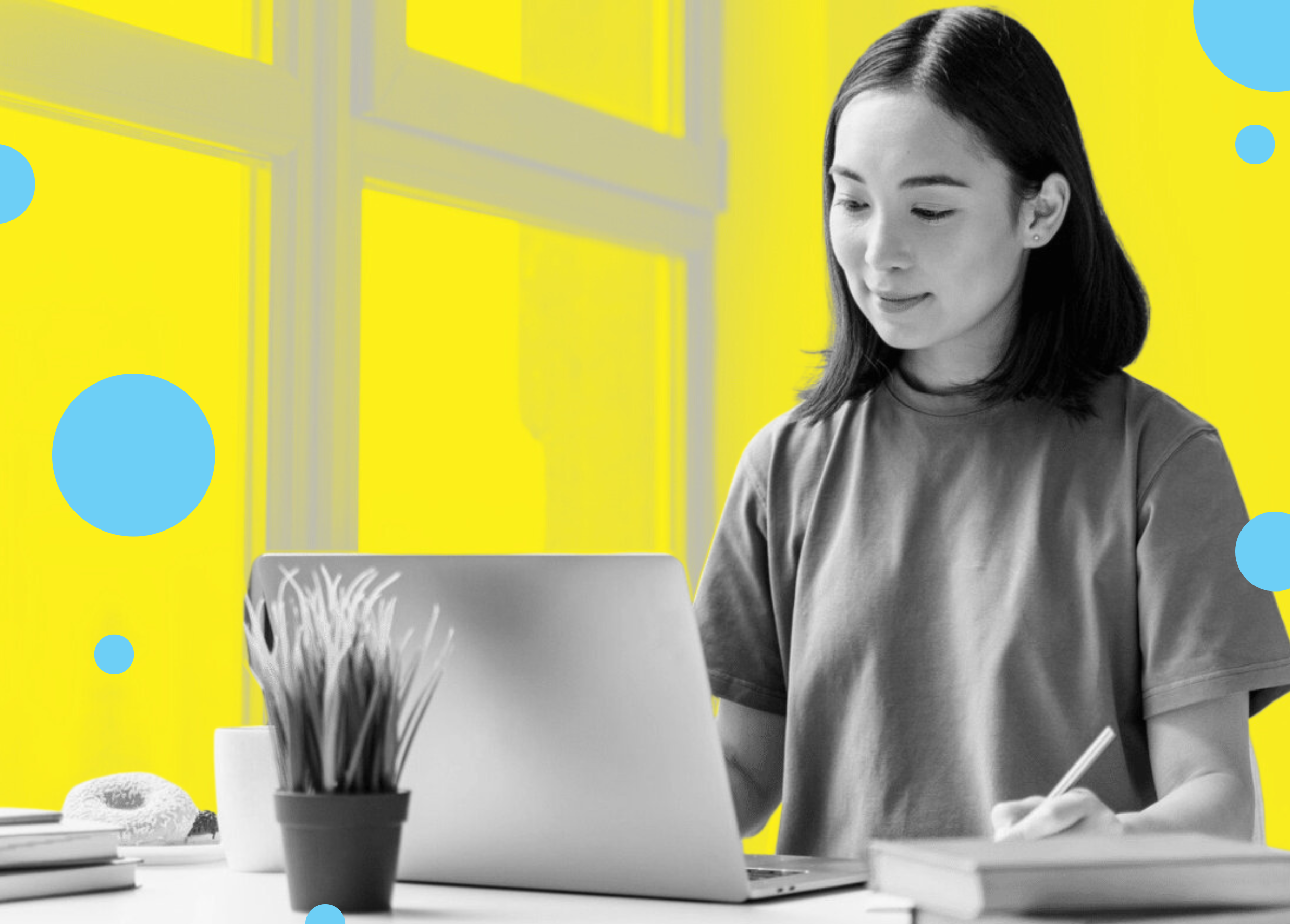 A person in a gray t-shirt is working on a laptop, writing notes with a pen, surrounded by a plant and a donut on the table.