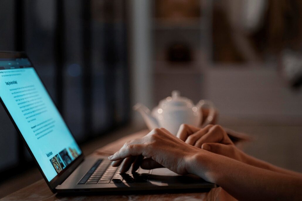 Hands typing on laptop with teapot in cozy workspace.