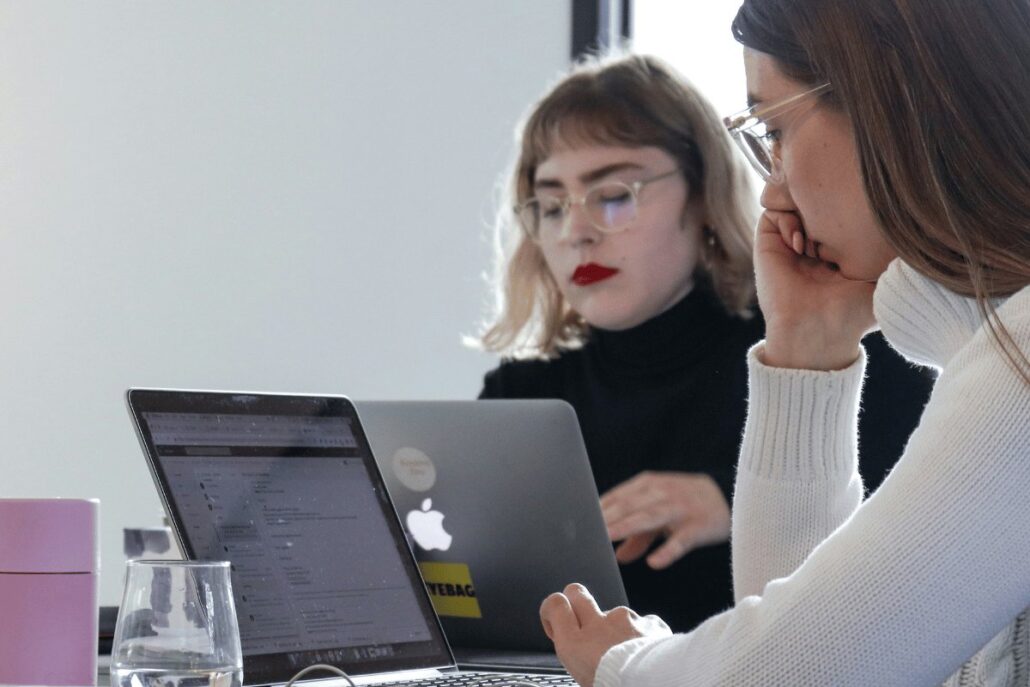 Two women working on laptops in a modern office setting.