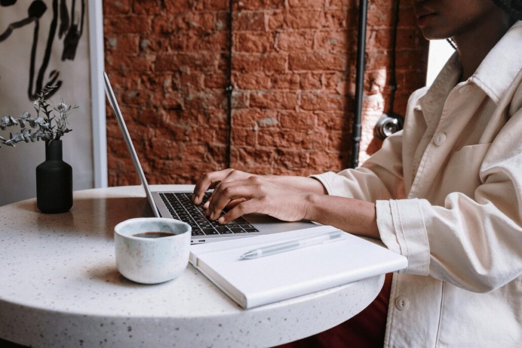 A person typing on a laptop at a café with a cup of coffee and a notebook on the table.