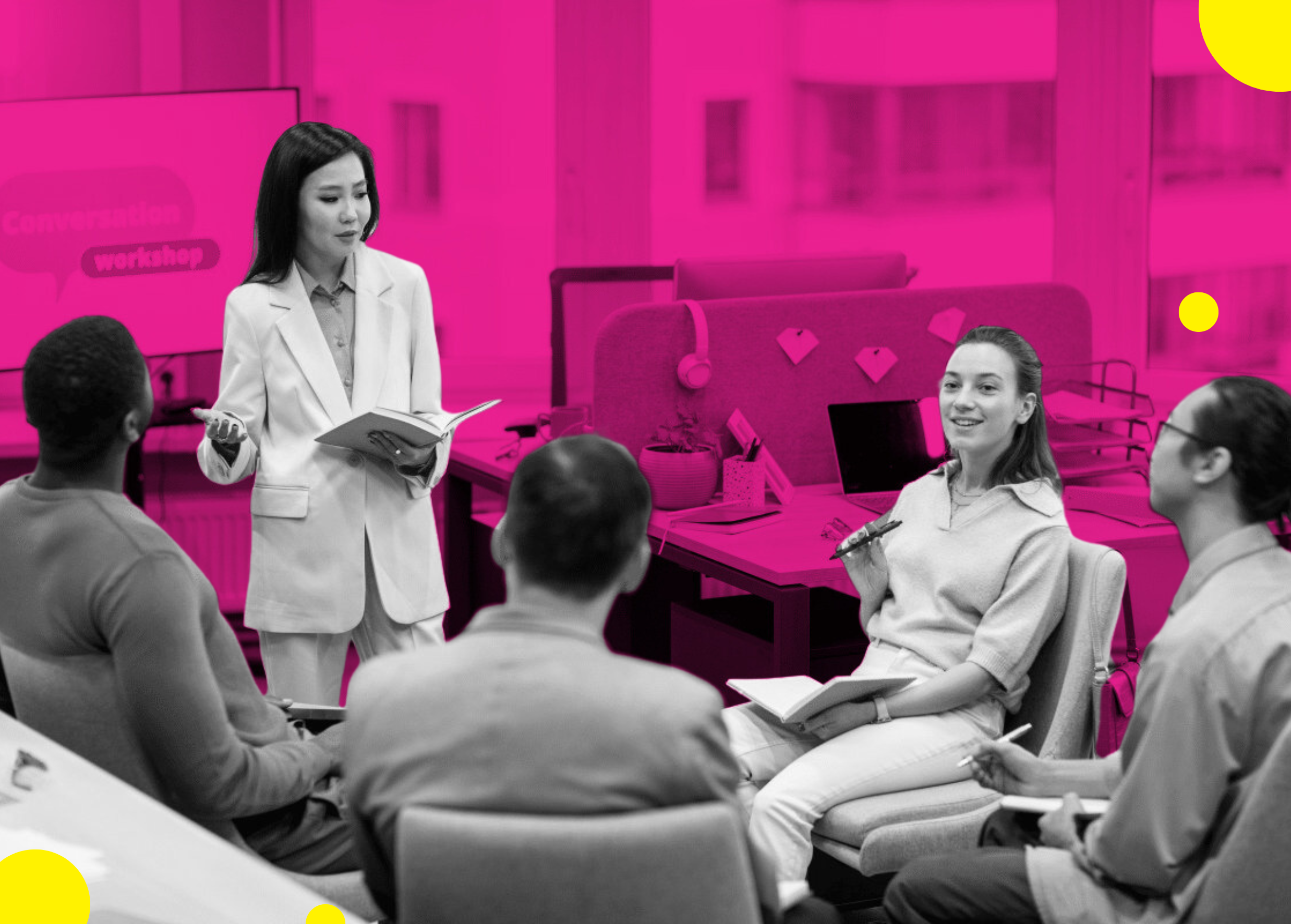 A woman in a white suit leads a discussion with a group seated in a modern office with bright pink accents. Participants appear engaged and focused.